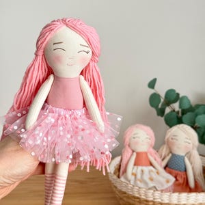 Puede incluir: Mu&ntilde;eca de tela hecha a mano con pelo de hilo rosa, un vestido rosa y una falda de tul con lunares rosas. Dos mu&ntilde;ecas m&aacute;s est&aacute;n en una cesta de mimbre en el fondo.