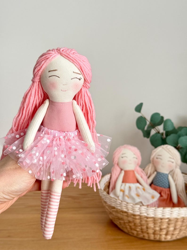Puede incluir: Mu&ntilde;eca de tela hecha a mano con pelo de hilo rosa, un vestido rosa y una falda de tul con lunares rosas. Dos mu&ntilde;ecas m&aacute;s est&aacute;n en una cesta de mimbre en el fondo.
