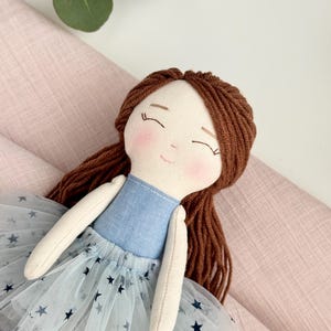 May include: A handmade doll with brown yarn hair and a light blue dress. The doll has a light blue bodice and a tulle skirt with dark blue star patterns. The doll's face has rosy cheeks and closed eyes.