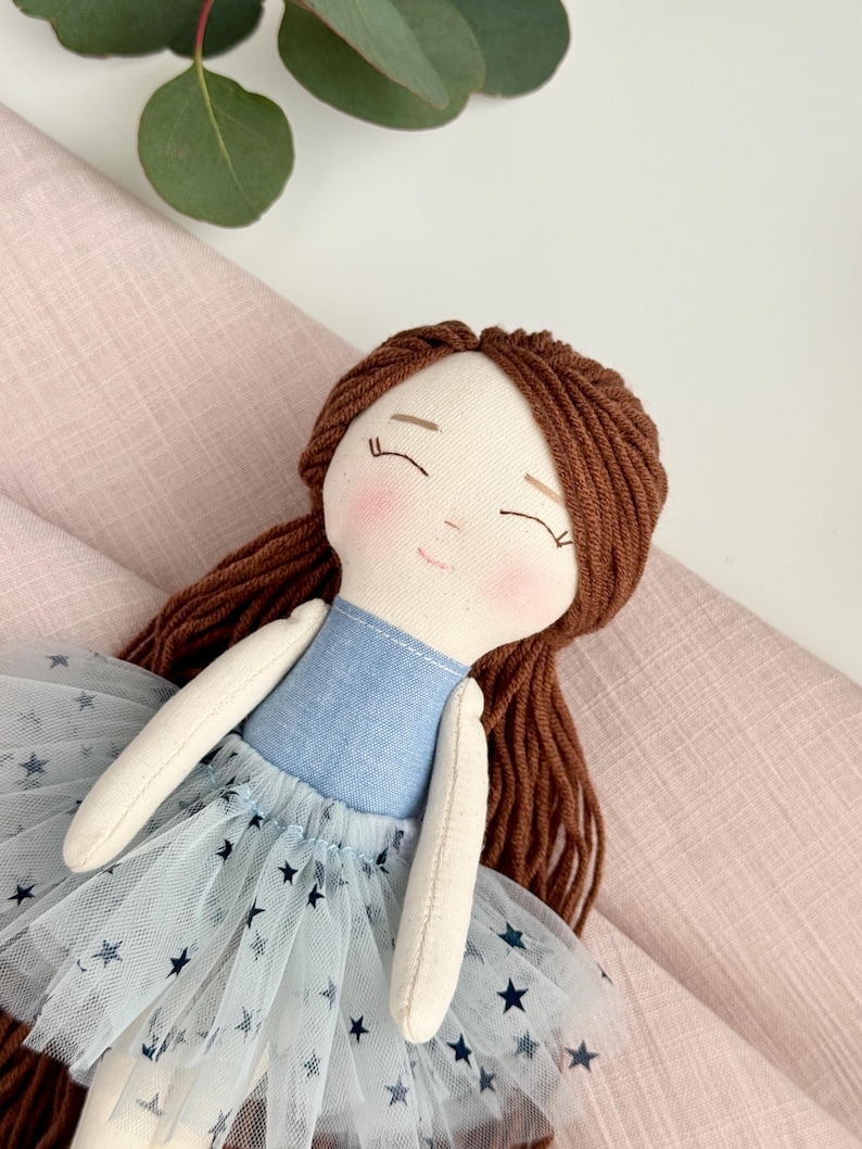 May include: A handmade doll with brown yarn hair and a light blue dress. The doll has a light blue bodice and a tulle skirt with dark blue star patterns. The doll's face has rosy cheeks and closed eyes.