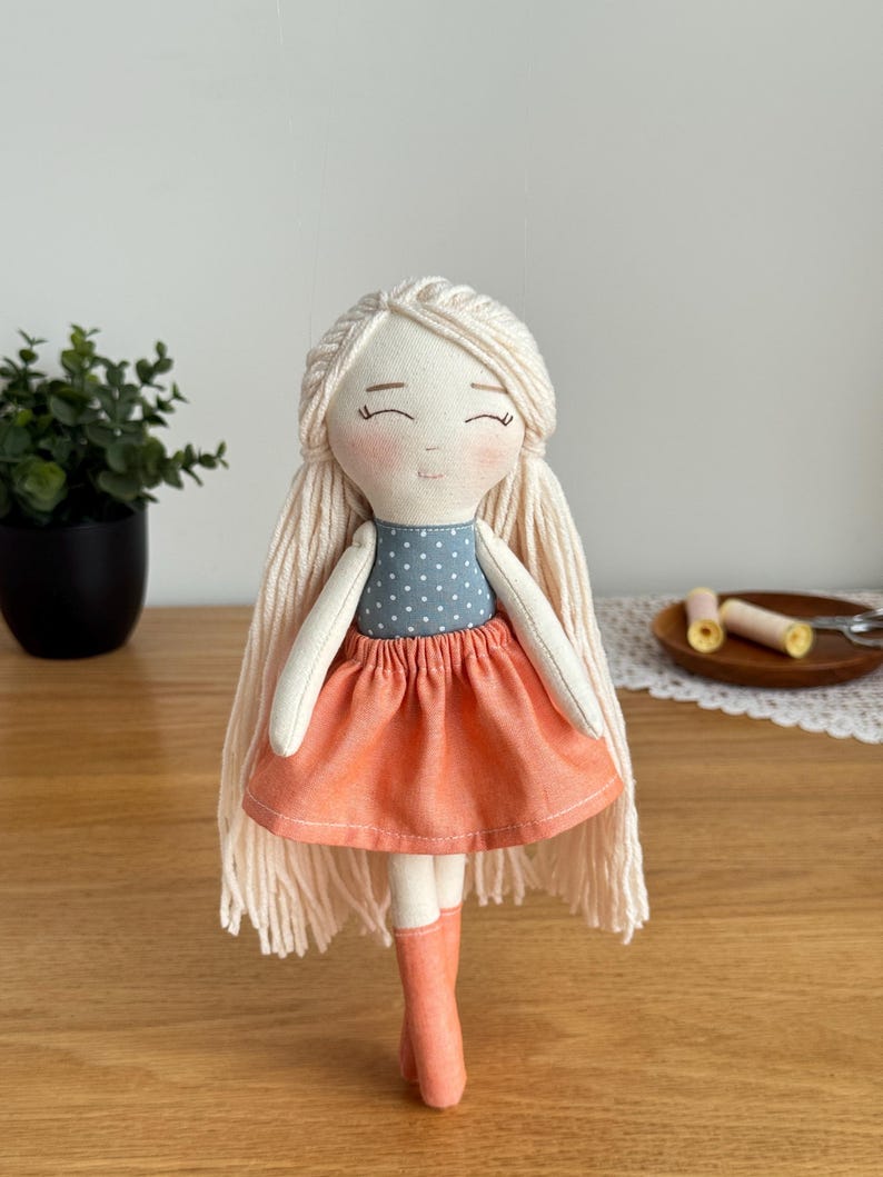 May include: A handmade fabric doll with long, light blonde yarn hair and a serene expression. The doll wears a blue polka-dot top, an orange skirt, and matching orange boots. The doll is a charming example of handcrafted textile art.