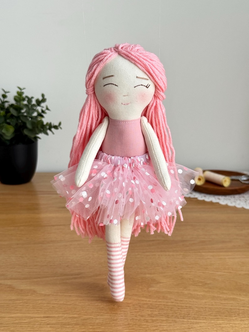 Puede incluir: Mu&ntilde;eca de tela hecha a mano con pelo de hilo rosa y una cara sonriente. La mu&ntilde;eca lleva un maillot rosa, un tut&uacute; con lunares blancos y calcetines a rayas. Un juguete encantador y caprichoso.