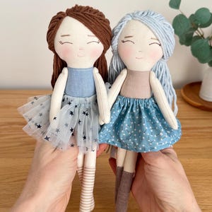 May include: Two handmade cloth dolls with smiling faces. One doll has brown yarn hair, a blue dress, and a tulle skirt with star accents. The other doll has blue yarn hair and a blue floral skirt. Both dolls have striped socks.