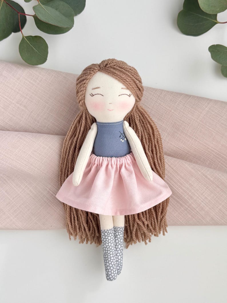 Peut inclure: Poup&eacute;e en tissu faite &agrave; la main avec de longs cheveux bruns en fil, un haut bleu et une jupe rose. La poup&eacute;e a un visage doux et souriant avec des joues roses et des chaussettes grises &agrave; motifs. Un article charmant et artisanal.