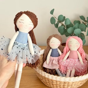 May include: Three handmade cloth dolls with yarn hair. One doll is held up, wearing a blue top and a tulle skirt with star accents. Two other dolls sit in a woven basket, one with brown hair and a pink skirt, the other with pink hair and a pink tulle skirt.