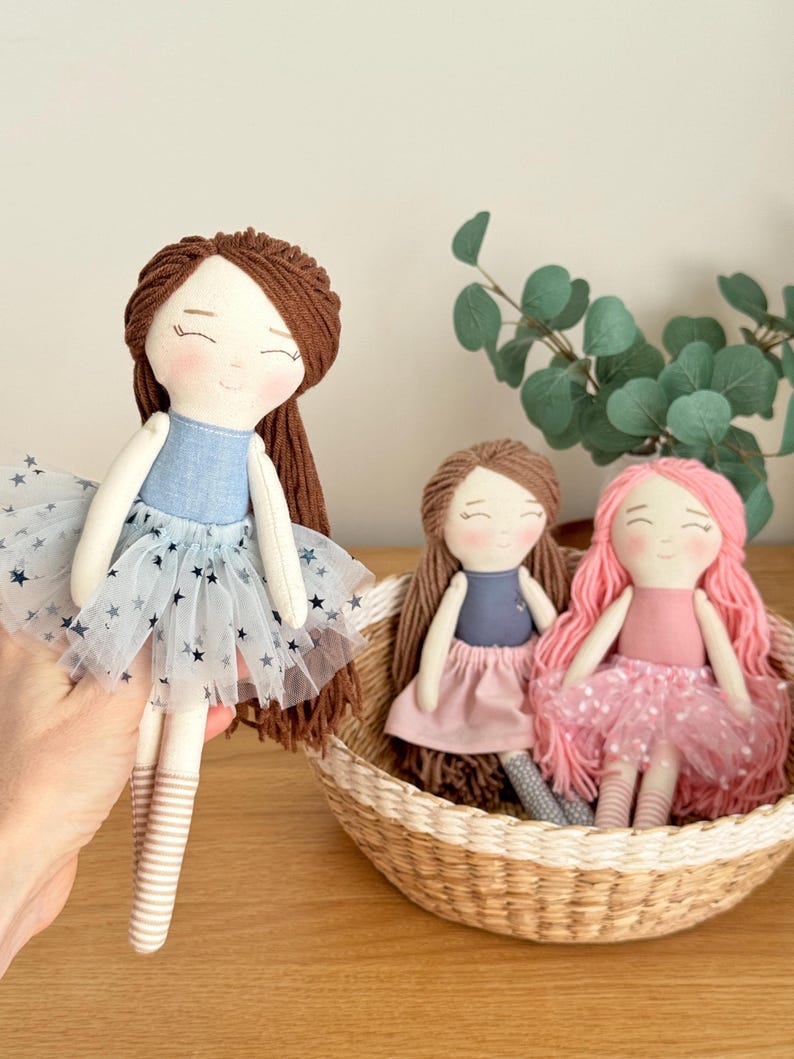 May include: Three handmade cloth dolls with yarn hair. One doll is held up, wearing a blue top and a tulle skirt with star accents. Two other dolls sit in a woven basket, one with brown hair and a pink skirt, the other with pink hair and a pink tulle skirt.