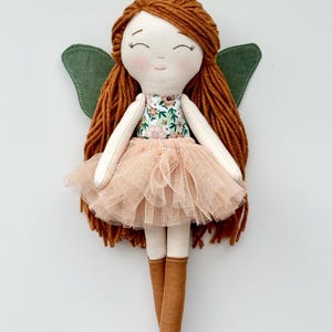 K&ouml;nnte beinhalten: Eine handgemachte Puppe mit langen, braunen Garnhaaren, einem Kleid mit Blumenmuster, einem T&uuml;llrock in Pfirsichfarbe, gr&uuml;nen Fl&uuml;geln und braunen Stoffschuhen.