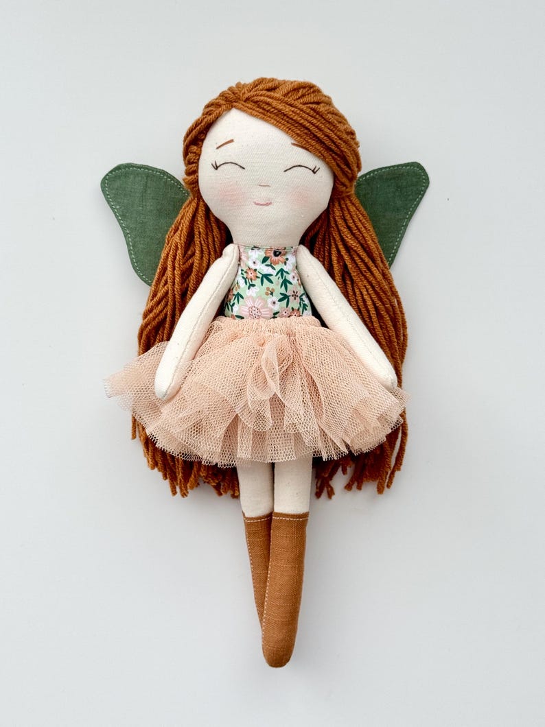K&ouml;nnte beinhalten: Eine handgemachte Puppe mit langen, braunen Garnhaaren, einem Kleid mit Blumenmuster, einem T&uuml;llrock in Pfirsichfarbe, gr&uuml;nen Fl&uuml;geln und braunen Stoffschuhen.
