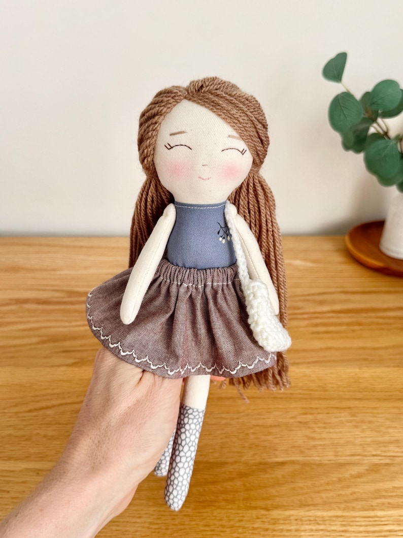 Peut inclure: Poup&eacute;e faite &agrave; la main avec des cheveux en fil marron et un haut bleu. La poup&eacute;e a une jupe marron avec un bord festonn&eacute; blanc et des leggings &agrave; motifs gris et blancs. Un petit sac &agrave; main blanc est suspendu &agrave; l'&eacute;paule de la poup&eacute;e.