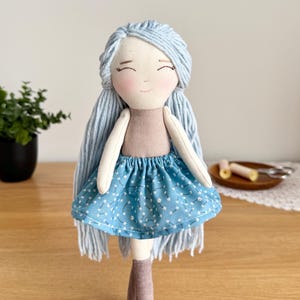 May include: A handmade doll with light blue yarn hair and a smiling face. The doll wears a tan top and a blue skirt with a white floral pattern. It has gray boots. The doll is a soft toy.