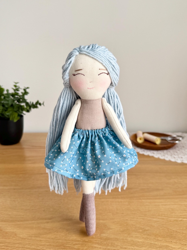 May include: A handmade doll with light blue yarn hair and a smiling face. The doll wears a tan top and a blue skirt with a white floral pattern. It has gray boots. The doll is a soft toy.