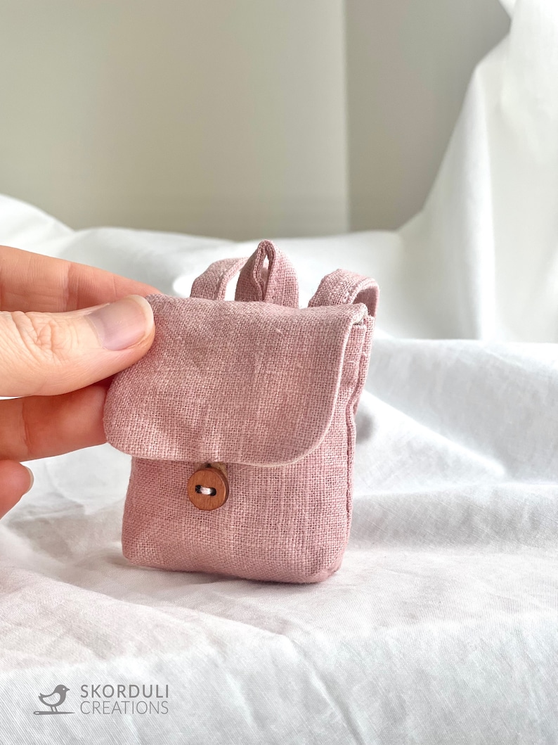May include: A small pink linen backpack with a wooden button closure. The backpack is made of a soft, textured fabric.