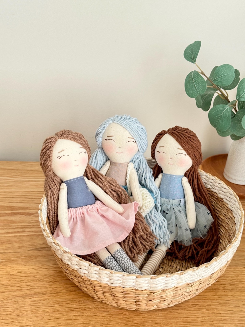 May include: Three handmade cloth dolls with yarn hair sit in a woven basket. The dolls have smiling faces and wear dresses in shades of blue and pink. One doll has a pink skirt, another has a blue skirt with star accents, and the third has a plain blue dress.