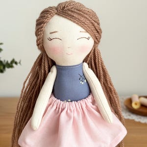 Peut inclure: Poup&eacute;e en tissu faite &agrave; la main avec des cheveux en fil brun et une expression sereine. La poup&eacute;e porte un haut bleu avec un motif floral et une jupe rose. Les bras et le visage de la poup&eacute;e sont beige clair.