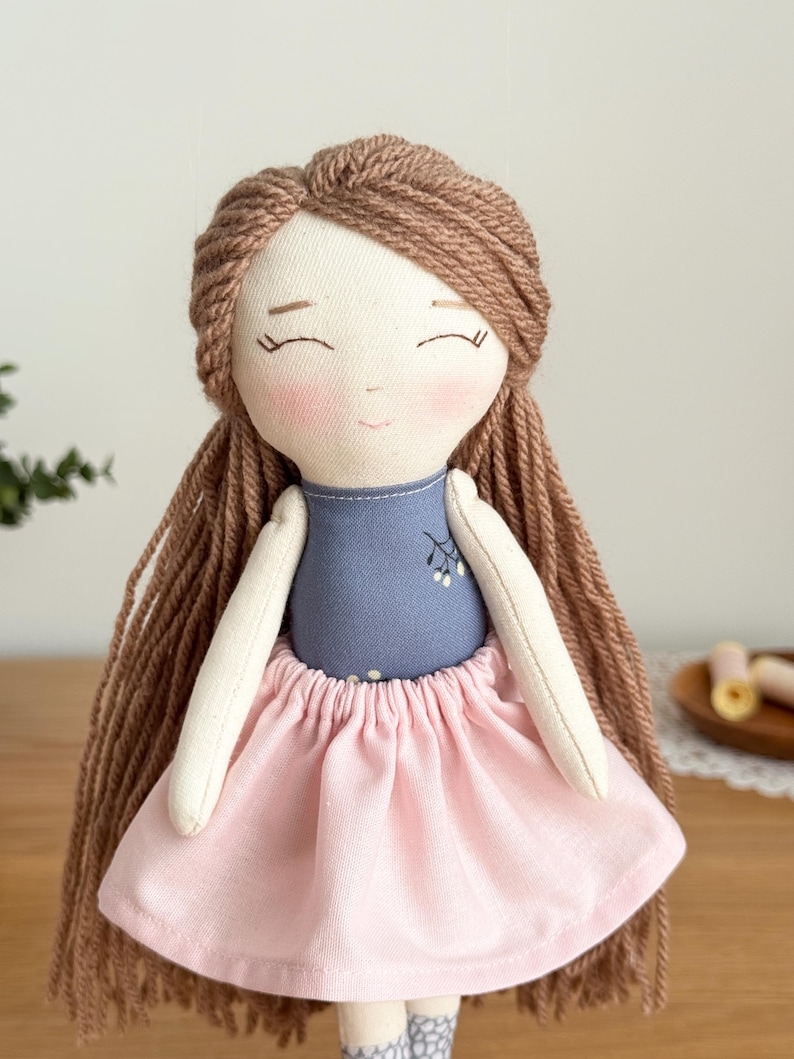 Peut inclure: Poup&eacute;e en tissu faite &agrave; la main avec des cheveux en fil brun et une expression sereine. La poup&eacute;e porte un haut bleu avec un motif floral et une jupe rose. Les bras et le visage de la poup&eacute;e sont beige clair.