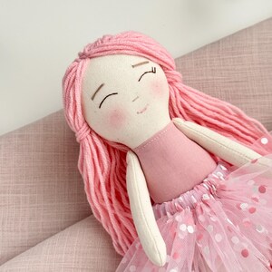 Puede incluir: Mu&ntilde;eca hecha a mano con pelo de hilo rosa y cuerpo de tela rosa. La mu&ntilde;eca tiene una falda tut&uacute; rosa con lunares blancos y rosas. La cara de la mu&ntilde;eca tiene los ojos cerrados y mejillas rosadas. Es un juguete blando.