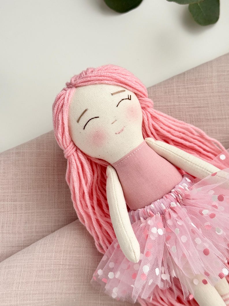 Puede incluir: Mu&ntilde;eca hecha a mano con pelo de hilo rosa y cuerpo de tela rosa. La mu&ntilde;eca tiene una falda tut&uacute; rosa con lunares blancos y rosas. La cara de la mu&ntilde;eca tiene los ojos cerrados y mejillas rosadas. Es un juguete blando.