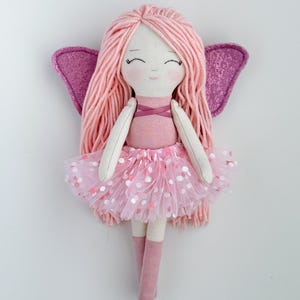 K&ouml;nnte beinhalten: Eine rosa Feenpuppe mit langen rosa Garnhaaren, einem rosa T&uuml;llrock mit wei&szlig;en Punkten und rosa Glitzerfl&uuml;geln.