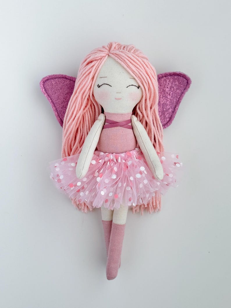 K&ouml;nnte beinhalten: Eine rosa Feenpuppe mit langen rosa Garnhaaren, einem rosa T&uuml;llrock mit wei&szlig;en Punkten und rosa Glitzerfl&uuml;geln.