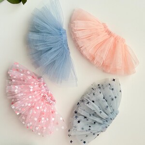 May include: Four miniature tutus in pastel colors. One is light blue, one is peach, one is pink with white and pink polka dots, and one is light blue with dark blue stars. The tutus are made of tulle.