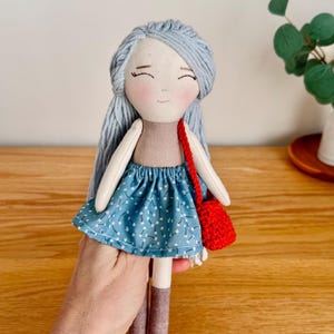 May include: Handmade doll with light blue yarn hair, a tan top, and a blue skirt with white floral accents. The doll has brown boots and a small red crocheted purse. The doll's face has a gentle expression.
