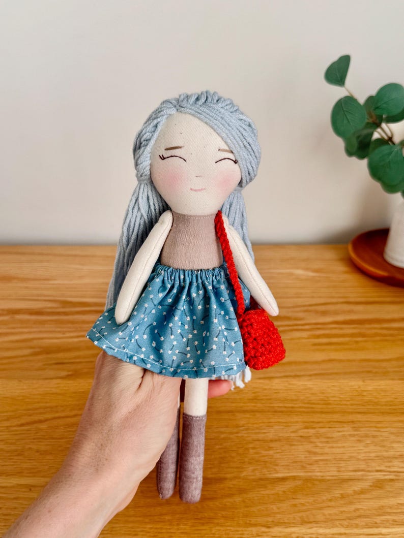 May include: Handmade doll with light blue yarn hair, a tan top, and a blue skirt with white floral accents. The doll has brown boots and a small red crocheted purse. The doll's face has a gentle expression.