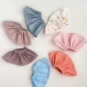 May include: A collection of seven fabric baby bonnets in various colors and patterns. The bonnets include solid colors like pink and coral, and patterned ones with white polka dots and small bird illustrations. Each bonnet has a gathered design.
