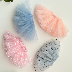 May include: Four miniature tutus in pastel colors. One is light blue, one is peach, one is pink with white and pink polka dots, and one is light blue with dark blue stars. The tutus are made of tulle.