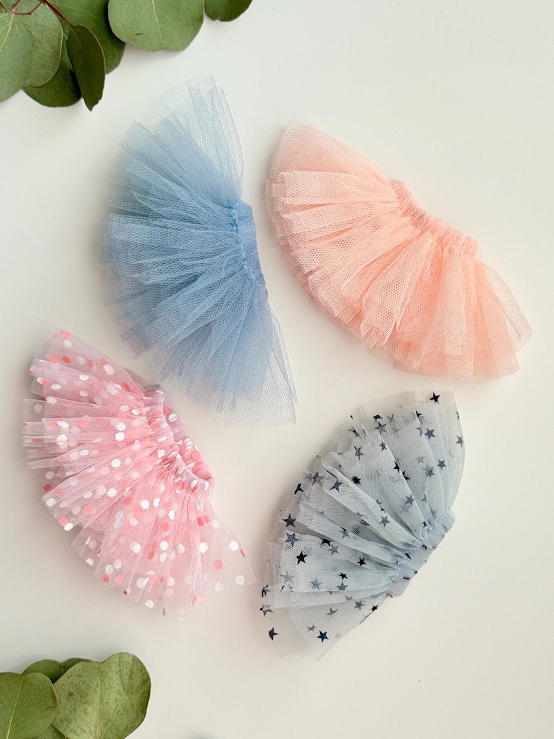 May include: Four miniature tutus in pastel colors. One is light blue, one is peach, one is pink with white and pink polka dots, and one is light blue with dark blue stars. The tutus are made of tulle.