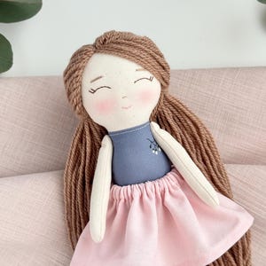 Peut inclure: Une poup&eacute;e faite &agrave; la main avec des cheveux en fil brun clair, des joues roses et les yeux ferm&eacute;s. La poup&eacute;e porte un haut bleu avec un motif floral et une jupe rose. Les bras et les jambes de la poup&eacute;e sont en tissu cr&egrave;me.