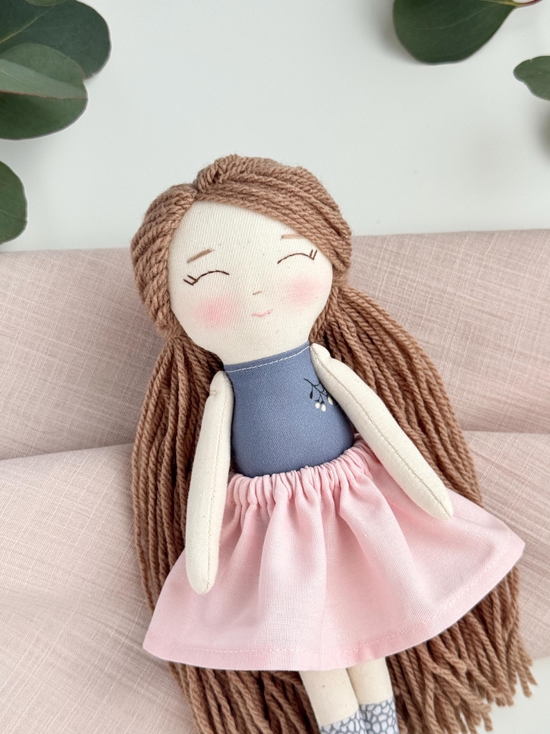 Peut inclure: Une poup&eacute;e faite &agrave; la main avec des cheveux en fil brun clair, des joues roses et les yeux ferm&eacute;s. La poup&eacute;e porte un haut bleu avec un motif floral et une jupe rose. Les bras et les jambes de la poup&eacute;e sont en tissu cr&egrave;me.