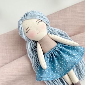 May include: A handmade doll with a light blue yarn hair, a tan top, and a blue skirt with white floral patterns. The doll has a smiling face with rosy cheeks and brown boots. The doll is lying on a pink fabric.