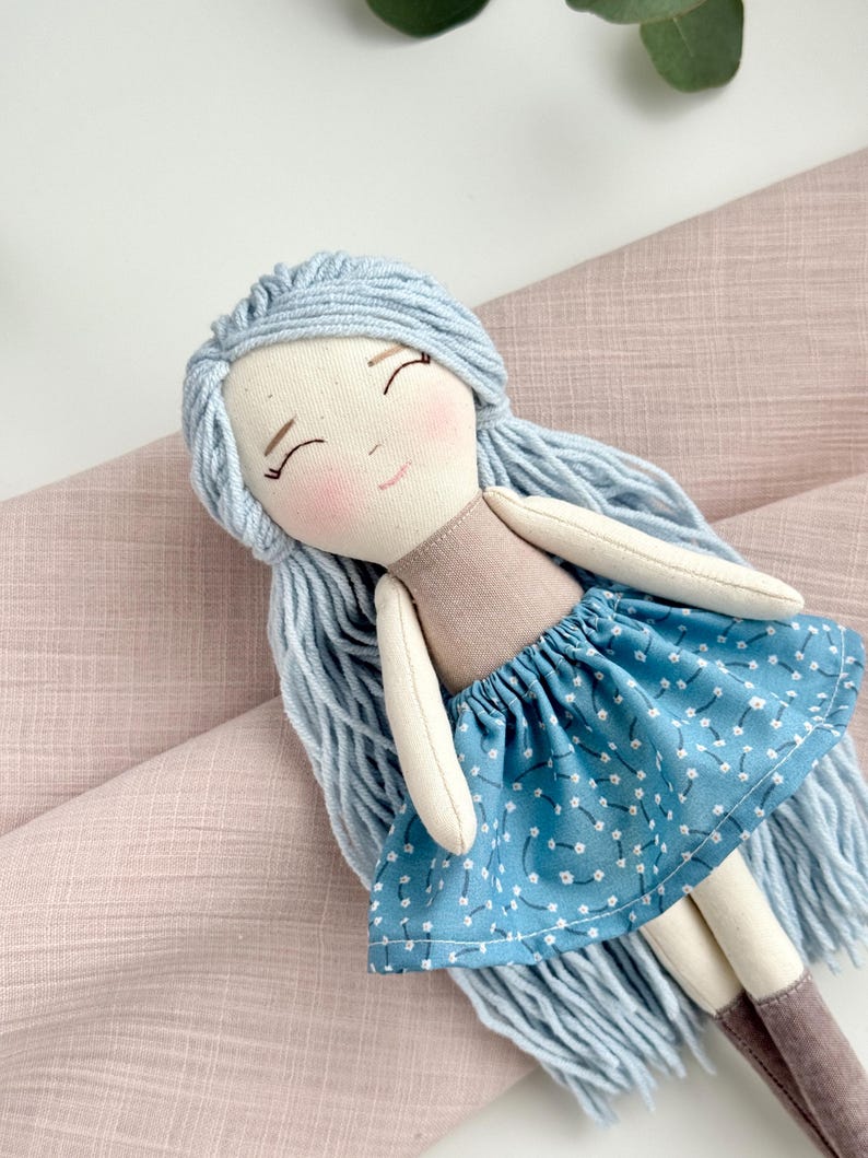 May include: A handmade doll with a light blue yarn hair, a tan top, and a blue skirt with white floral patterns. The doll has a smiling face with rosy cheeks and brown boots. The doll is lying on a pink fabric.