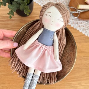 Peut inclure: Poup&eacute;e en tissu faite &agrave; la main avec des cheveux en fil marron, un haut bleu et une jupe rose. La poup&eacute;e a des joues roses et porte des chaussettes grises et blanches &agrave; motifs. La poup&eacute;e repose dans un bol en laiton d&eacute;coratif.