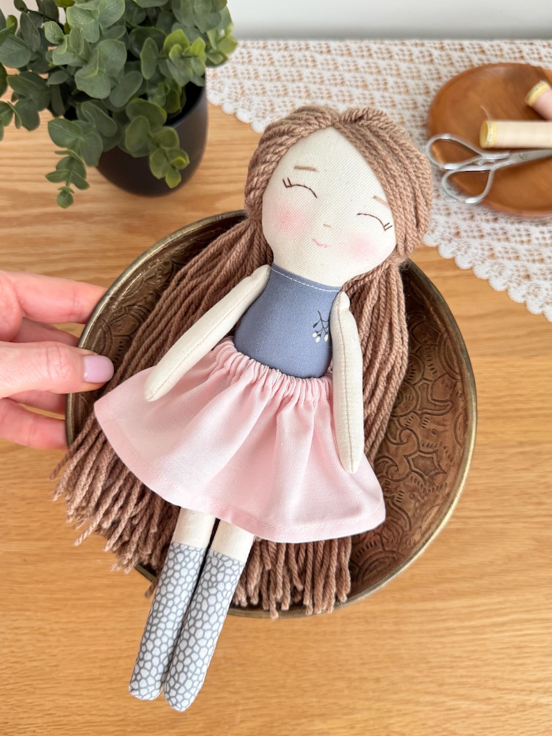 Peut inclure: Poup&eacute;e en tissu faite &agrave; la main avec des cheveux en fil marron, un haut bleu et une jupe rose. La poup&eacute;e a des joues roses et porte des chaussettes grises et blanches &agrave; motifs. La poup&eacute;e repose dans un bol en laiton d&eacute;coratif.