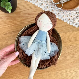 May include: A handmade doll with brown yarn hair, a blue bodice, and a light blue tulle skirt with star accents. The doll has striped legs and is resting in a decorative brass bowl. The doll's face has a sweet, hand-stitched expression.