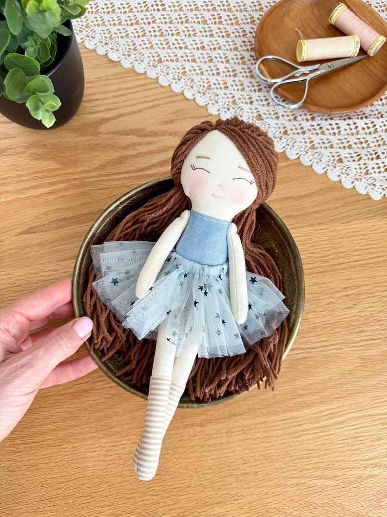 May include: A handmade doll with brown yarn hair, a blue bodice, and a light blue tulle skirt with star accents. The doll has striped legs and is resting in a decorative brass bowl. The doll's face has a sweet, hand-stitched expression.