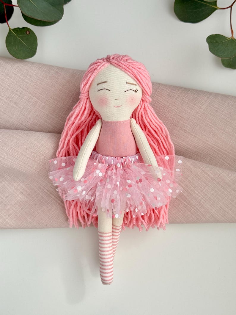 Puede incluir: Mu&ntilde;eca de tela hecha a mano con pelo de hilo rosa, un vestido rosa claro y un tut&uacute; rosa con lunares blancos y rosas. La mu&ntilde;eca tiene una cara sonriente y calcetines a rayas. Es un juguete infantil encantador.