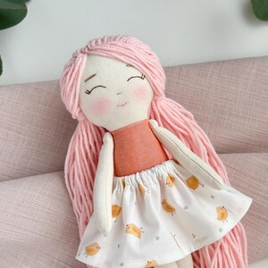 May include: A handmade fabric doll with pink yarn hair and rosy cheeks. The doll wears a peach-colored top and a white skirt printed with yellow birds. The doll's arms and legs are made of a cream-colored fabric.