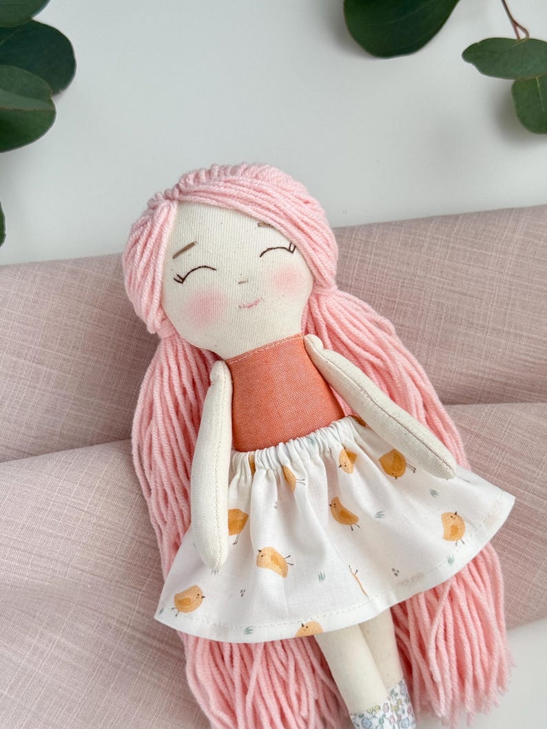 May include: A handmade fabric doll with pink yarn hair and rosy cheeks. The doll wears a peach-colored top and a white skirt printed with yellow birds. The doll's arms and legs are made of a cream-colored fabric.
