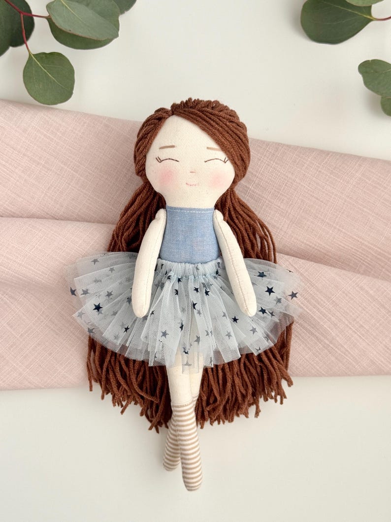 May include: A handmade doll with brown yarn hair, a blue denim bodice, and a white tulle skirt adorned with navy blue stars. The doll has a sweet, smiling face and is wearing striped socks. A charming, handcrafted toy.