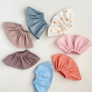 May include: A collection of seven fabric baby bonnets in various colors and patterns. The bonnets include solid colors like pink and coral, and patterned ones with white polka dots and small bird illustrations. Each bonnet has a gathered design.