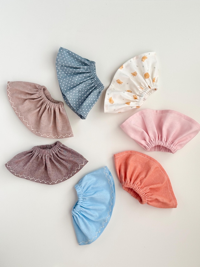 May include: A collection of seven fabric baby bonnets in various colors and patterns. The bonnets include solid colors like pink and coral, and patterned ones with white polka dots and small bird illustrations. Each bonnet has a gathered design.