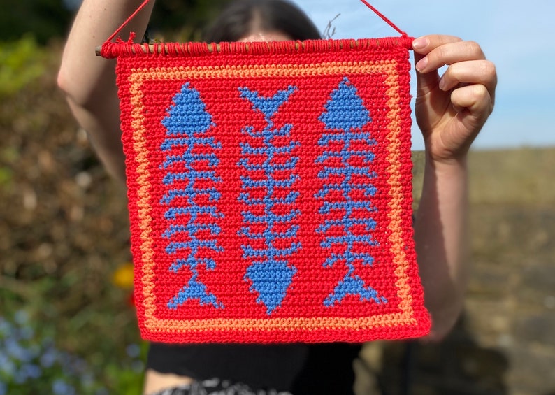 May include: A red crocheted wall hanging with a design of three blue fish skeletons. The hanging has a peach colored border.