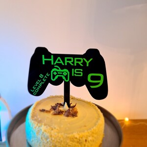May include: Black and green gamer-themed cake topper with the text "HARRY IS 9" and a green video game controller icon. The topper also includes the text "LEVEL 8 COMPLETE" on the side.