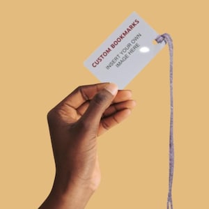 May include: A white bookmark with the text "CUSTOM BOOKMARKS" and "INSERT YOUR OWN IMAGE HERE" in red. The bookmark is held by a hand and has a purple string attached. The background is a solid, muted orange.