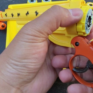 May include: A yellow and orange toy gun with a removable cylinder. The cylinder has a black and white sticker with the word "RELOAD" on it. The gun is being held by a hand.