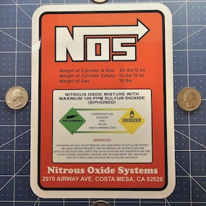 Puede incluir: Una pegatina rectangular con un diseño rojo y blanco, con el texto "NOS" e información sobre el óxido nitroso. La pegatina incluye detalles de peso en libras y onzas, junto con advertencias de seguridad y la dirección de Nitrous Oxide Systems.