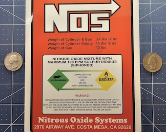 Puede incluir: Una pegatina rectangular naranja y blanca de Nitrous Oxide Systems con el logotipo NOS, especificaciones de peso en lbs y oz, y advertencias de seguridad. La etiqueta incluye símbolos de peligro para gas no inflamable y oxidante.