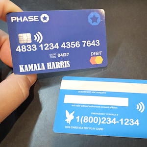 May include: Two blue toy credit cards are displayed. The front card has the Chase logo, a contactless symbol, and the name "KAMALA HARRIS". The card number is 4833 1234 4356 7643, with an expiration date of 04/27. The back card has a contactless symbol and the text "EMERGENCY CONTACT # 1(800)234-1234".